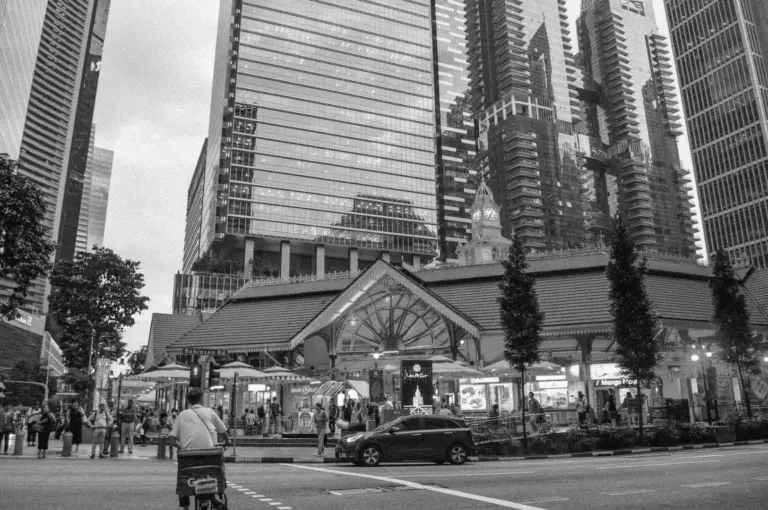 Black and white photo of a bustling city intersection. A market with a pitched roof is surrounded by modern skyscrapers, creating a blend of old and new.