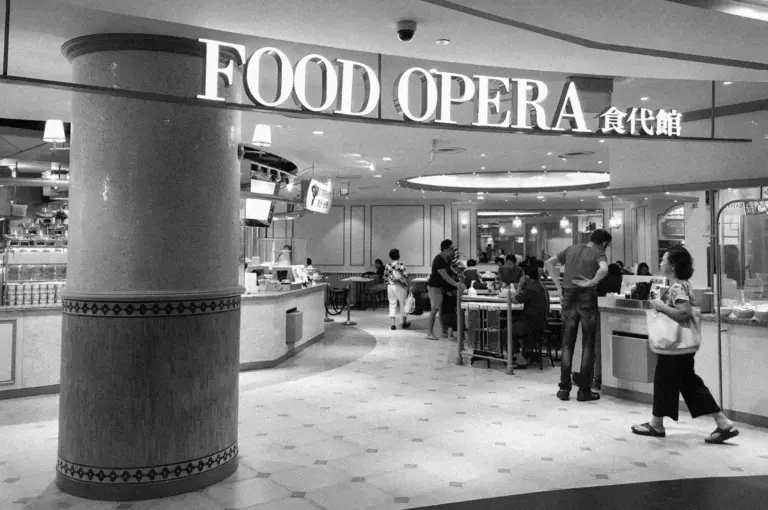 Black and white photo of a food court entrance labeled "Food Opera." People are dining and conversing, creating a lively and social atmosphere.