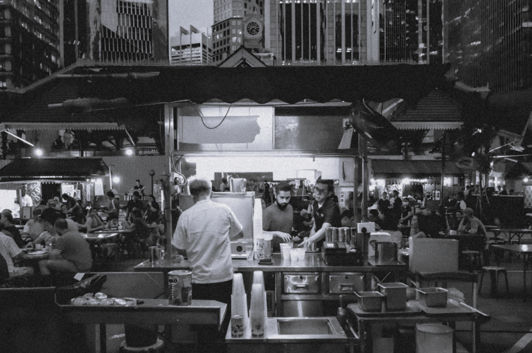 A bustling outdoor food court at dusk with a drink stall in the foreground, where two workers serve beverages. Diners fill tables under city skyscrapers.