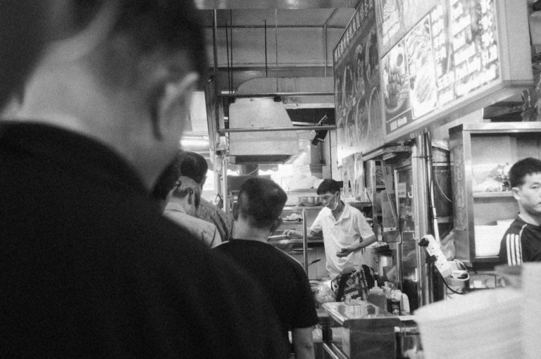 Black and white photo of a bustling street food stall with customers in line. A staff member prepares food under menu signs, evoking a lively, busy atmosphere.
