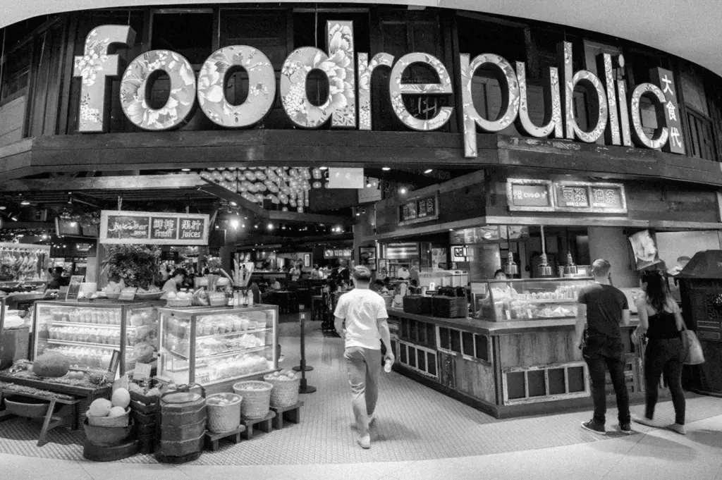Black and white image of a bustling food court entrance labeled "food republic." Shoppers walk in, past colorful displays, exuding a lively, inviting vibe.