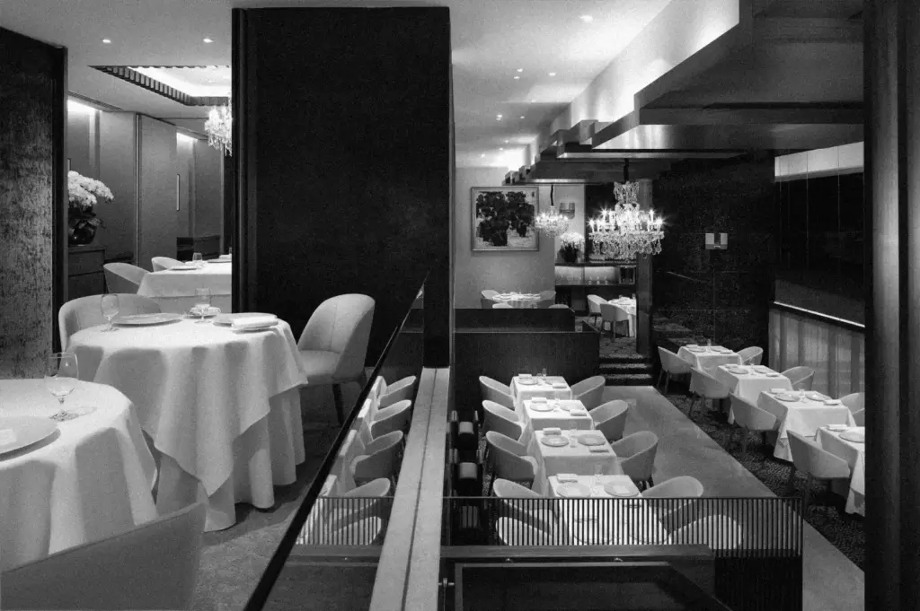 Elegant restaurant interior in black and white, featuring round and rectangular tables with white tablecloths, chandeliers, and modern decor. Cozy ambiance.