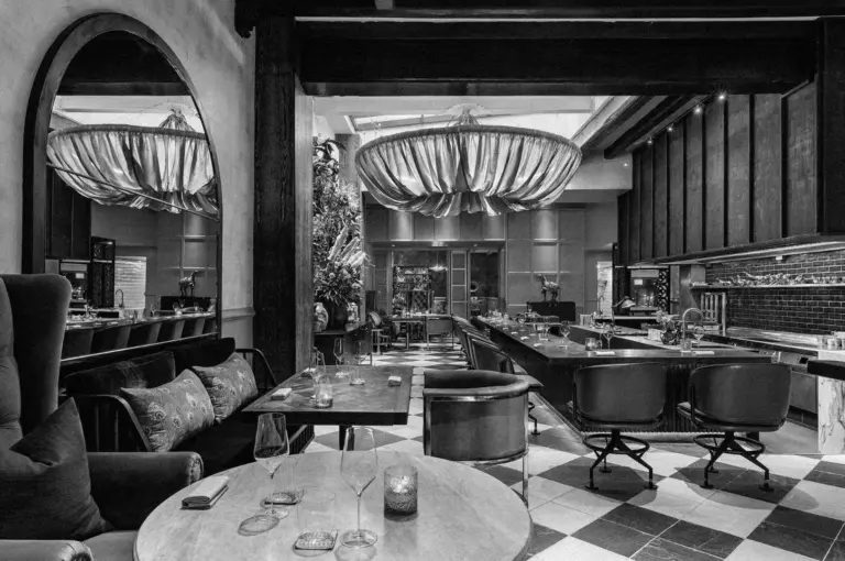 Chic modern bar in grayscale with plush seating and large mirrors. A circular chandelier hangs above, creating a cozy, stylish ambiance.