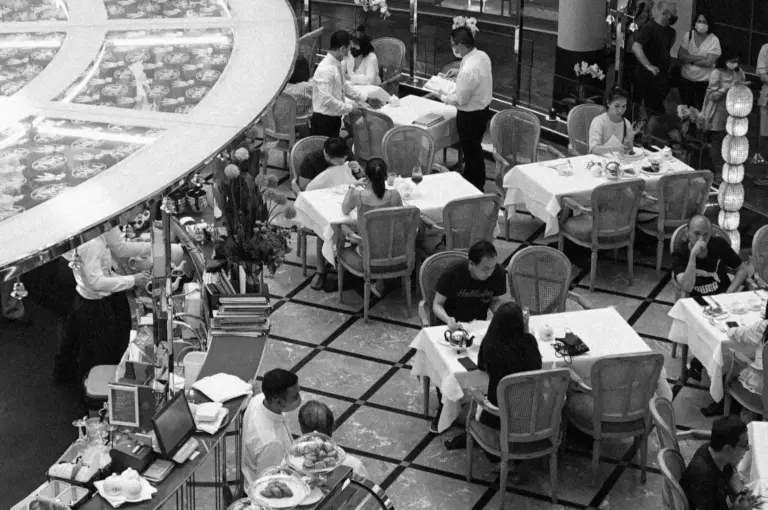Black and white image of a busy restaurant scene, with waitstaff serving patrons at tables set with white linens. The atmosphere is lively and bustling.