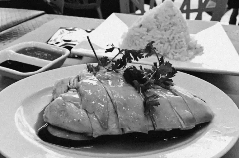 Plated dish of sliced chicken with a glossy sauce, garnished with cilantro. Background shows a pyramid of rice. Accompanied by small dishes of sauce.