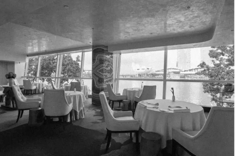 Elegant restaurant interior with round tables and white chairs, overlooking a city waterfront through large windows. Peaceful and luxurious ambiance.