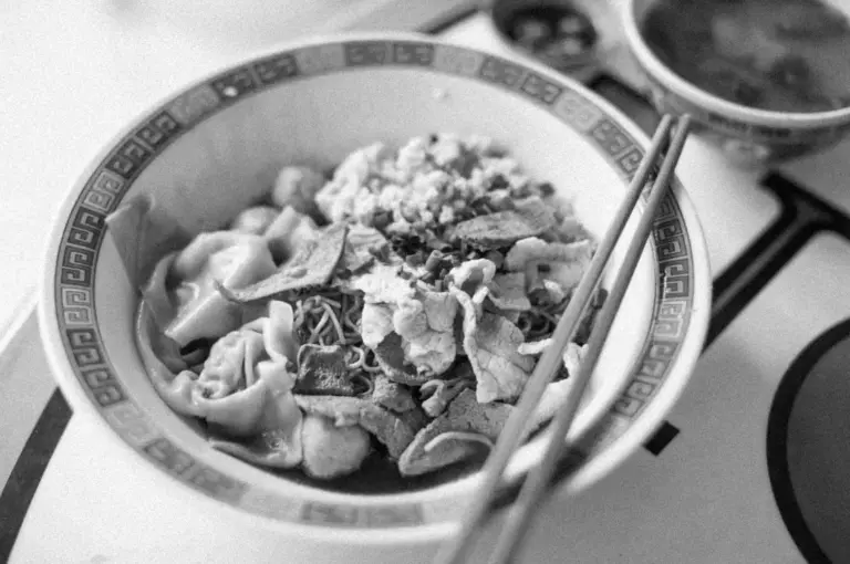 A black and white image of a bowl containing noodles, dumplings, and crispy toppings. Chopsticks rest on the bowl, with a small soup dish nearby.
