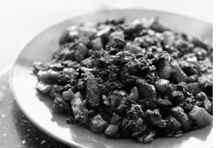 Black and white image of a plate filled with cubes of roasted meat and vegetables, creating a rustic and hearty presentation. Shadows add depth.