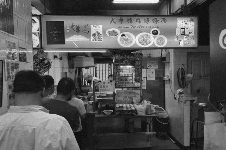 Black and white image of a bustling noodle stall with a Michelin 2023 sign. Three people in line, staff preparing food behind a counter with various dishes displayed above. Inviting and busy atmosphere.