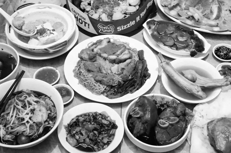 A variety of Asian dishes displayed on a table, including noodles, soup, fish, and vegetables. The composition conveys a sense of abundance and richness.