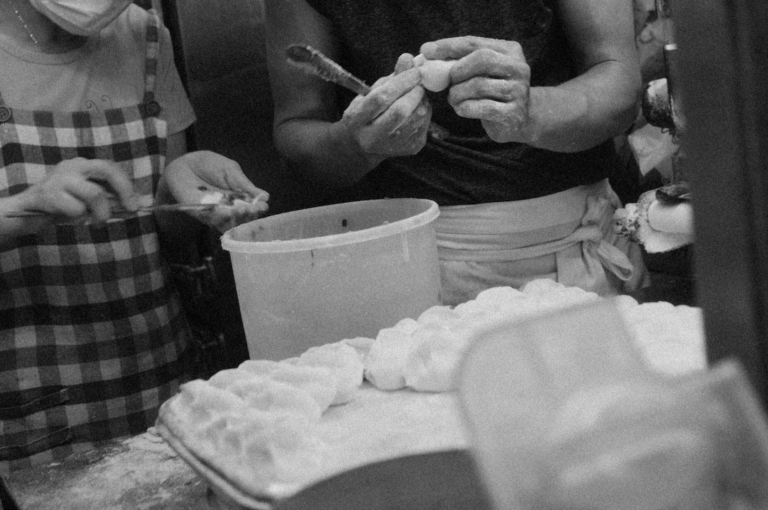 Black and white photo of two people making dumplings. One holds a dumpling with tongs, while the other shapes dough on a flour-dusted tray.