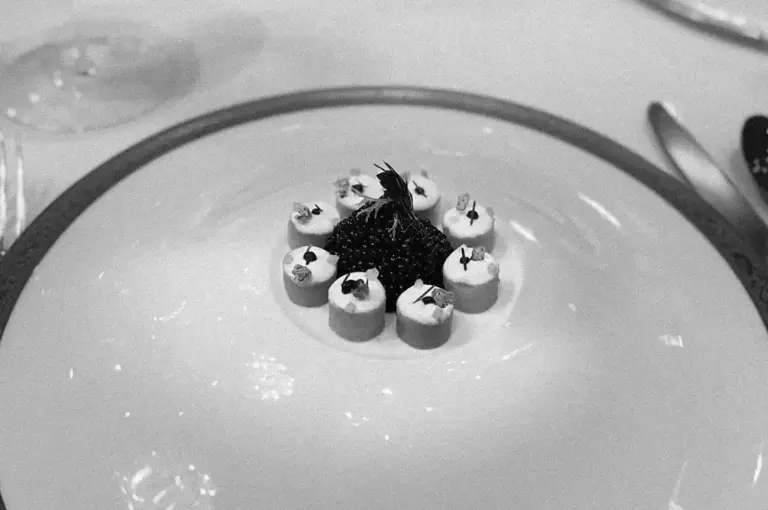 A monochrome image of a gourmet dish featuring a central mound of caviar surrounded by neatly arranged small dollops of cream, garnished with herbs.
