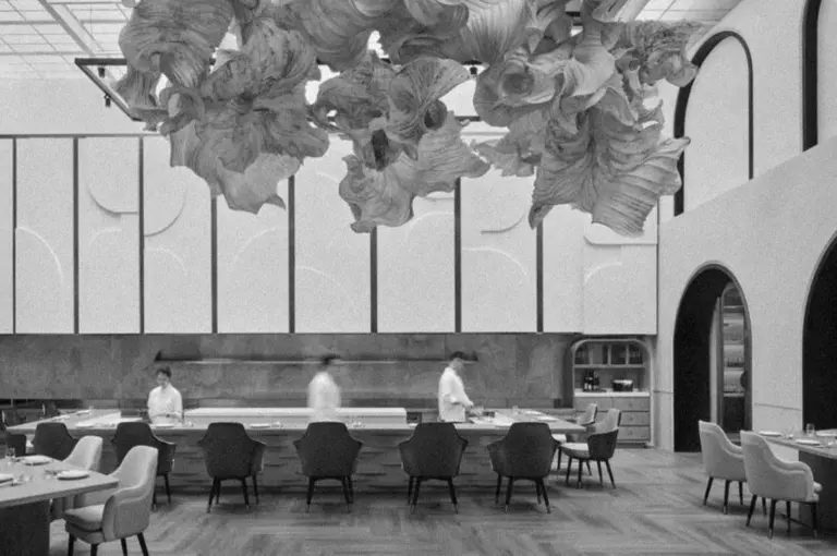 A modern restaurant interior with abstract sculptural ceiling art. Three chefs move behind a curved bar, surrounded by stylish chairs. Monochrome, elegant.