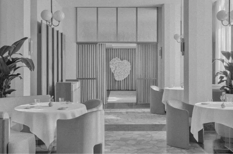 Black and white image of a modern, minimalistic restaurant with round tables, white tablecloths, and elegant chairs. A heart-shaped decoration adds charm.