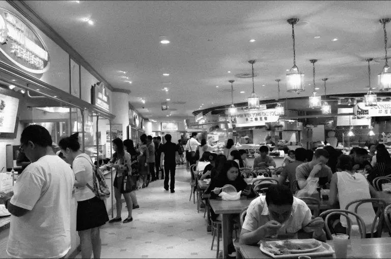 Black and white image of a bustling food court. People are lined up at counters on the left, while others sit and eat at tables on the right, creating a lively atmosphere.