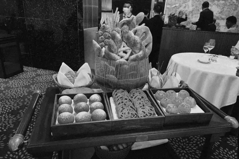 A black-and-white image of a restaurant bread cart with various artisanal breads, rolls, and baguettes in baskets. Elegant dining tables are visible in the background.