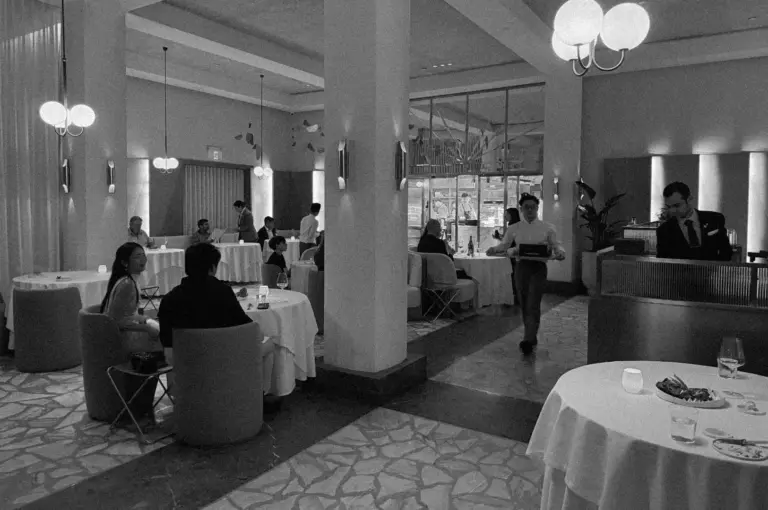 A dimly lit, elegant restaurant with diners at round tables, a waiter carrying a tray, and soft, ambient lighting. The atmosphere is calm and sophisticated.