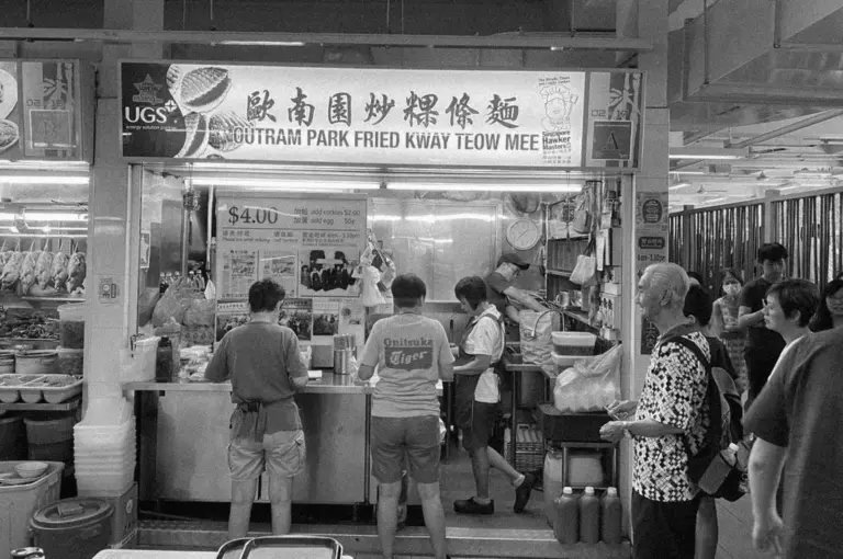 Black and white photo of a busy food stall named "Outram Park Fried Kway Teow Mee." Customers queue as a cook prepares food in an energetic scene.