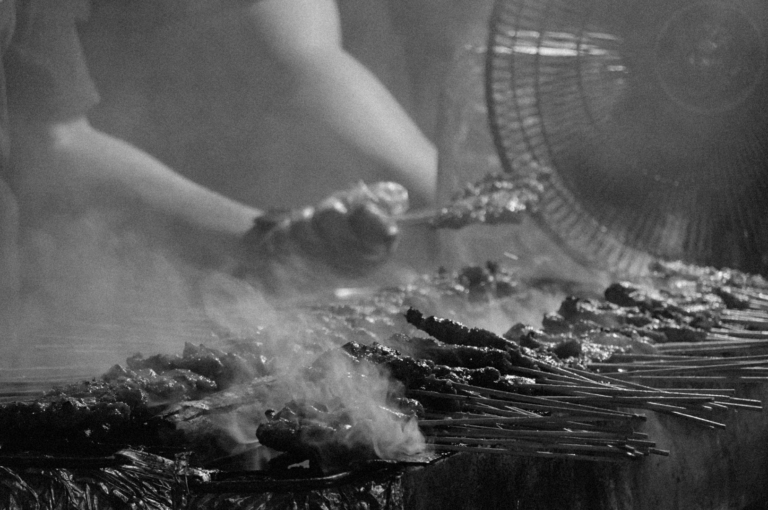 A person grills skewered meats over a smoky barbecue. A fan in the background disperses the smoke, creating a rustic, lively street food scene.