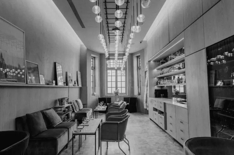 Black and white image of a modern, elegant lounge with plush sofas, a long table, and a stylish ceiling light fixture. Large windows add natural light.
