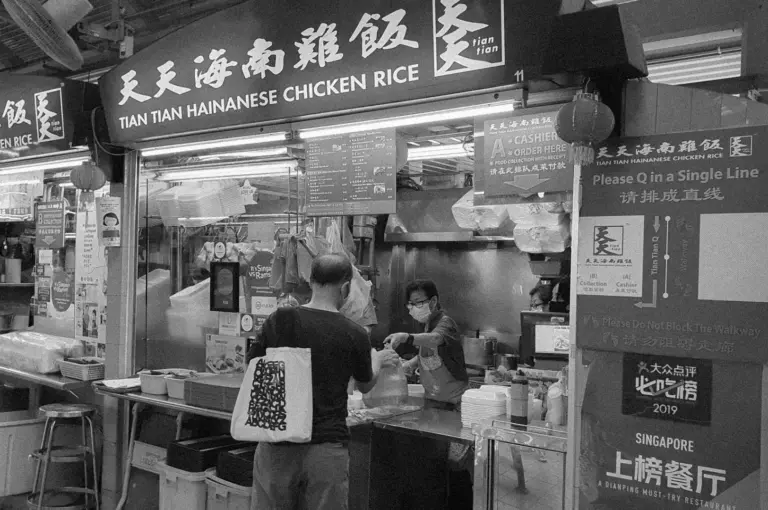 Black-and-white photo of a street food stall, "Tian Tian Hainanese Chicken Rice." A masked vendor serves a customer with a tote bag, conveying busy, traditional vibes. Signs in English and Chinese direct queuing.