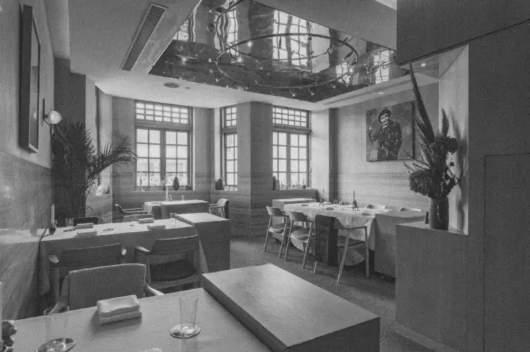 Modern restaurant interior in black and white, featuring neatly set tables, elegant chairs, large windows, a mirrored ceiling, and wall art, creating a sophisticated ambiance.