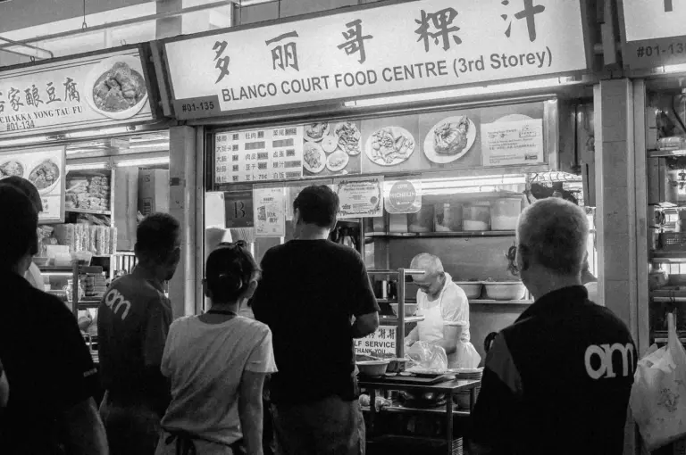 Black and white photo of a busy food stall at Blanco Court Food Centre. A small crowd waits as a vendor prepares dishes with concentration.