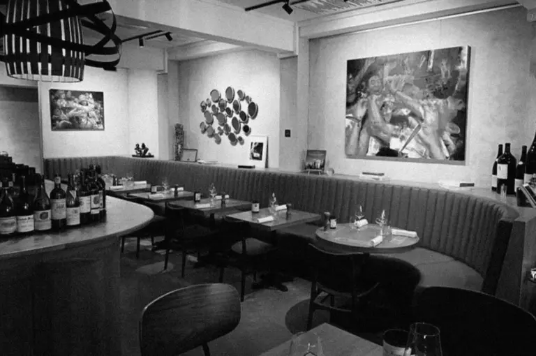 Cozy restaurant interior with round tables, cushioned booth seating, and contemporary art on walls. Wine bottles and dim lighting create an intimate atmosphere.