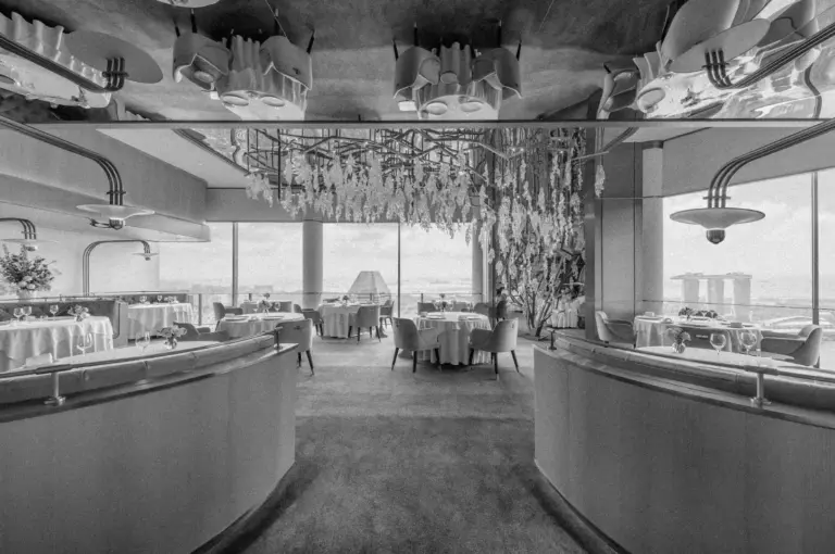 A black-and-white photo of an elegant, modern restaurant interior with a calm ambiance. Tables are set with white tablecloths under decorative lights, and large windows offer city views.