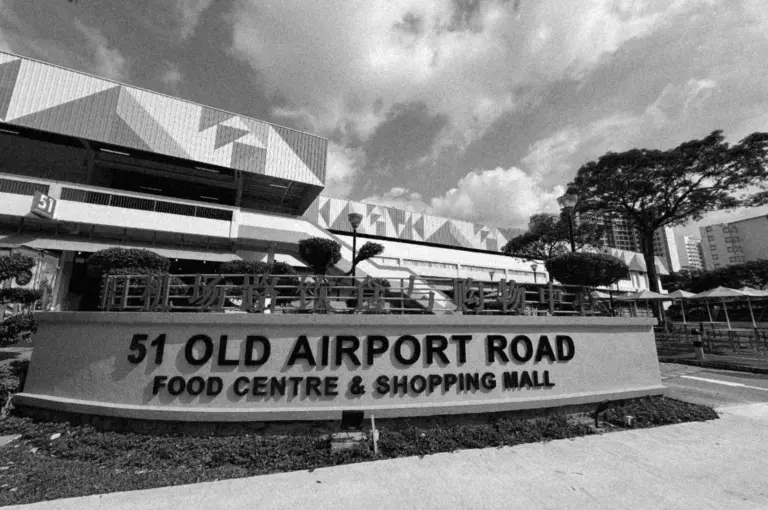 Black and white photo of 51 Old Airport Road Food Centre & Shopping Mall sign, modern building with geometric patterns, cloudy sky, and trees.