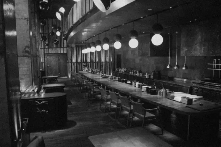 Dimly lit, empty restaurant interior with a long bar, wooden chairs, and hanging spherical lights. The atmosphere is modern and elegant.