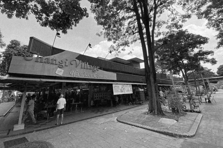 Black and white photo of Changi Village Hawker Centre. Features a tree-lined entrance with people gathered. Skies are partly cloudy. Relaxed vibe.