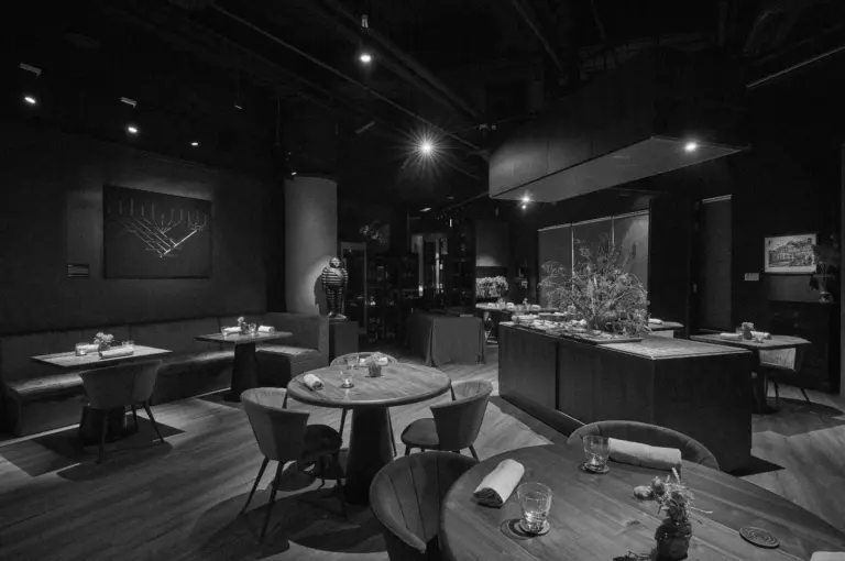 Dimly lit, elegant restaurant interior in black and white. Round wooden tables set with glasses and napkins. Modern, cozy ambiance with soft lighting.
