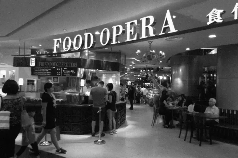 Black and white image of a bustling food court named "Food Opera." Patrons queue at a stall labeled "Fried Dough Fritters," with diners seated nearby.