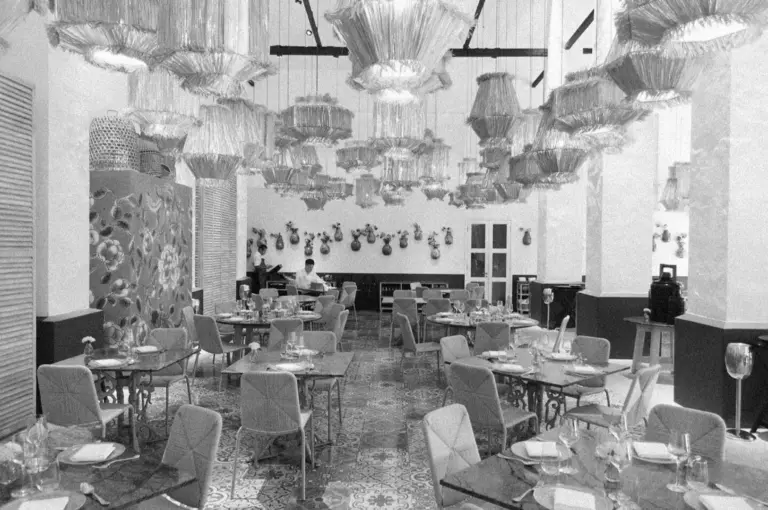 Black and white image of an elegant restaurant, featuring ornate hanging lights and neatly set tables with intricate patterns. The atmosphere is sophisticated and inviting.