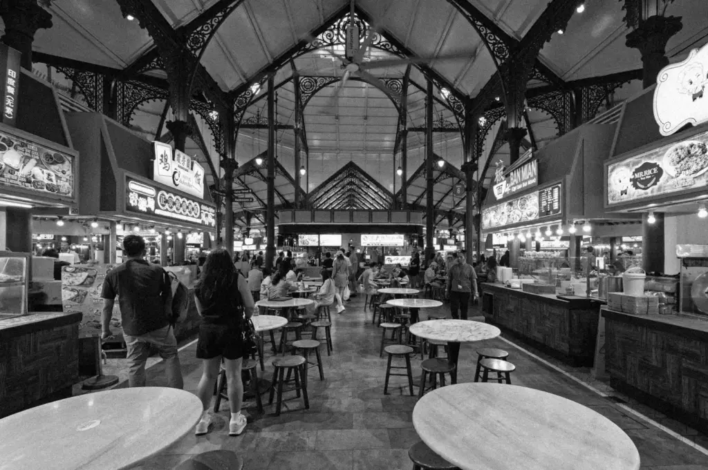 A bustling food hall with intricate ironwork and high ceilings. Empty tables in the foreground; food stalls and people create a lively atmosphere. Black and white tones