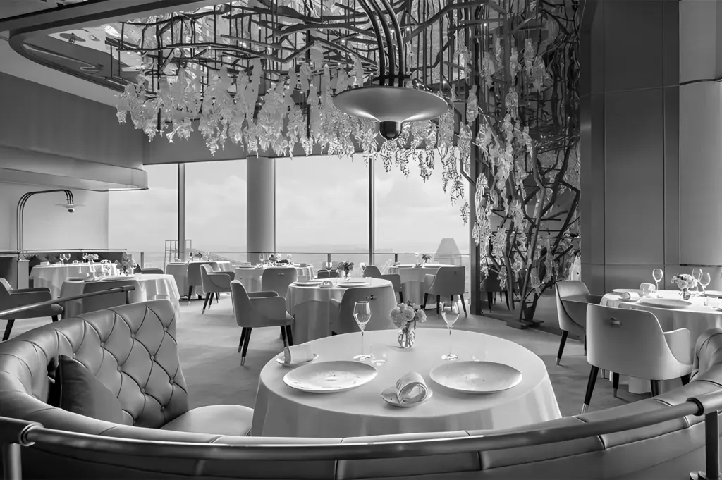 Elegant restaurant interior in black and white, featuring round tables with white tablecloths, plush chairs, and intricate hanging decorations. Minimalist and sophisticated ambiance.