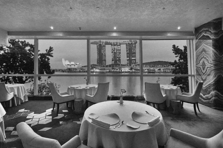 Elegant restaurant interior with round, white-clothed tables and cushioned chairs overlooking a scenic view of a city waterfront and modern skyscrapers.