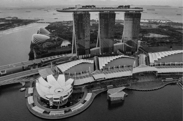 Aerial view of Marina Bay Sands in Singapore against a backdrop of water and ships. The iconic building features three towers and a rooftop garden, with the ArtScience Museum in the foreground. The tone is dynamic and architectural