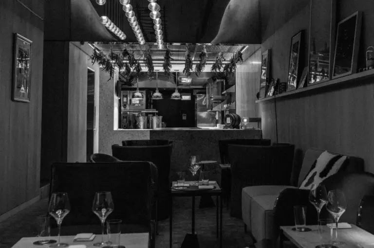 A modern restaurant interior in black and white, featuring cozy seating, dim lighting, a visible kitchen, and artworks on the walls, creating a relaxed, elegant atmosphere.