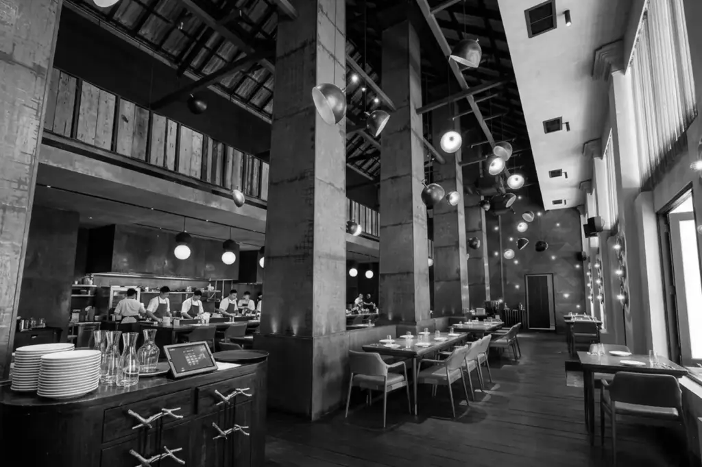 Spacious Burnt Ends dining room with high ceilings, concrete pillars, industrial pendant lights, and open kitchen visible in background