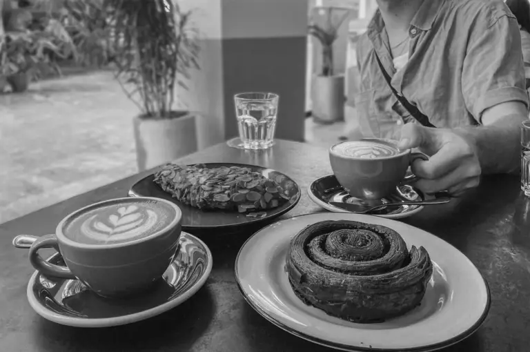 A cozy cafe scene with two lattes in green cups, a chocolate pastry, and a seed-covered treat on plates. A person in a blue shirt holds a cup.