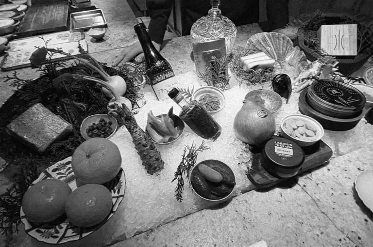 A monochrome image of a lavish table spread, featuring an assortment of fruits, vegetables, condiments, and ornate jars, exuding an elegant, gourmet ambiance.