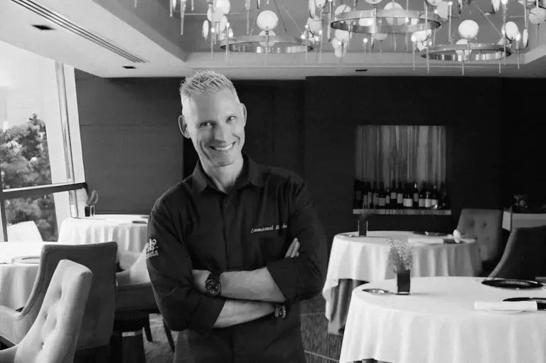 Smiling man with short hair stands confidently in an elegant restaurant, arms crossed. Tables are set, and modern chandeliers hang from the ceiling. Black and white.