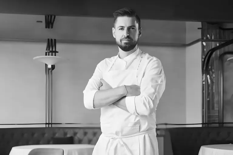 Chef in a crisp white uniform stands confidently with arms crossed in an elegant restaurant setting. The monochrome tone exudes professionalism and calm.