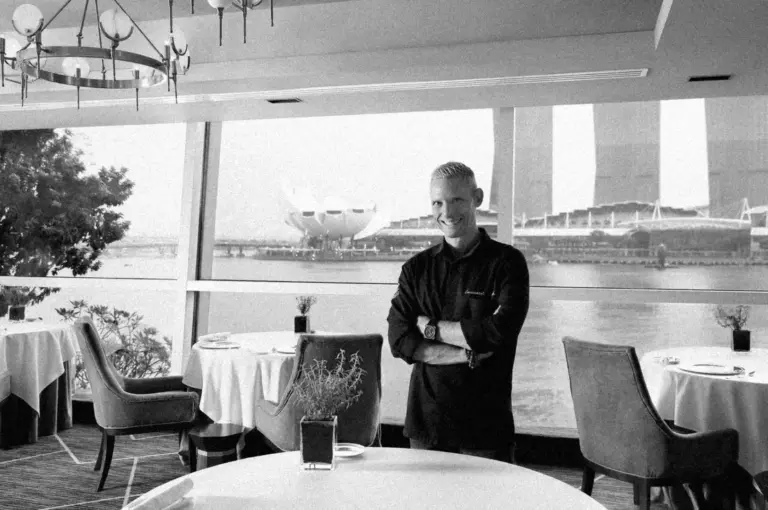 A smiling chef Emmanuel Stroobant stands with arms crossed in an elegant restaurant overlooking a waterfront. Large windows reveal iconic city landmarks outside.