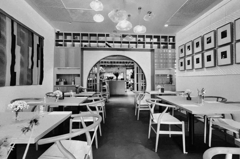 Black and white image of a modern, empty restaurant with elegant wooden chairs and tables. Art adorns the walls, with a softly lit, inviting ambiance