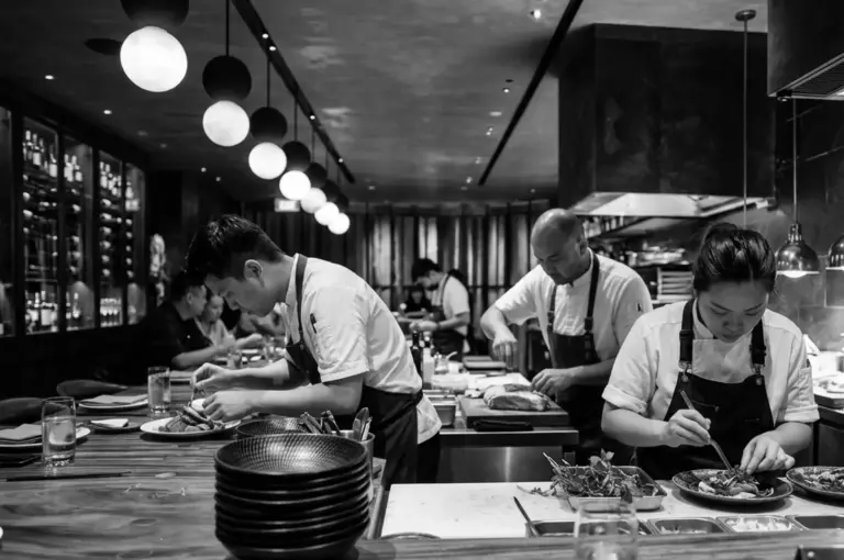 Chefs in aprons plating dishes at open kitchen counter with pendant lights and wine cellar in background