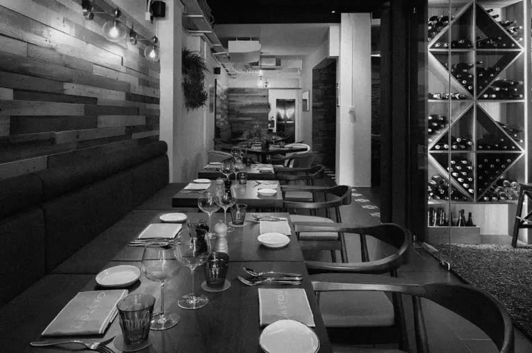 Black and white image of an elegant restaurant interior with neatly set tables, wooden paneling, and a wine rack, creating a cozy, sophisticated ambiance