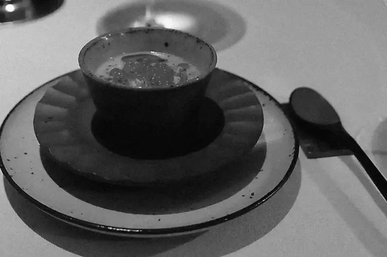 A black and white image of a dark, textured bowl on two stacked plates, with a spoon on the right. The setting is elegant and minimalistic, evoking a refined dining atmosphere.